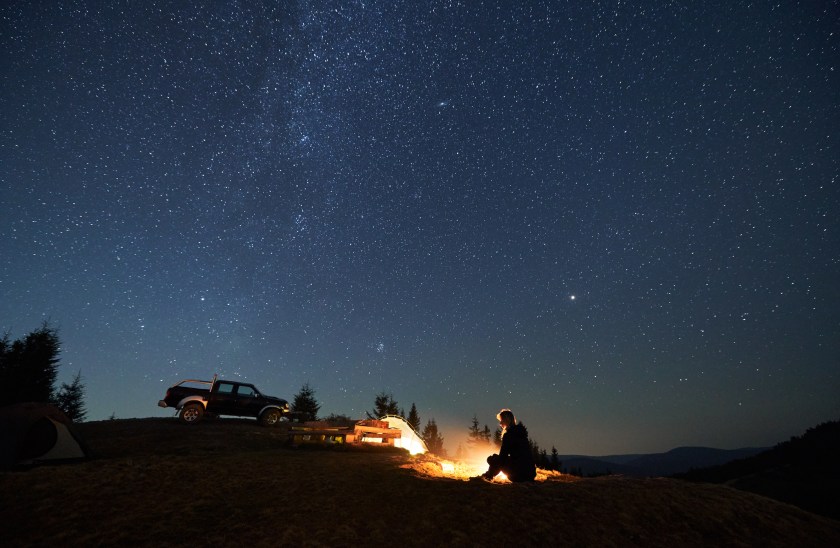 Car camper resting by bonfire, enjoying a starry night sky with her tent set up and her car close by.