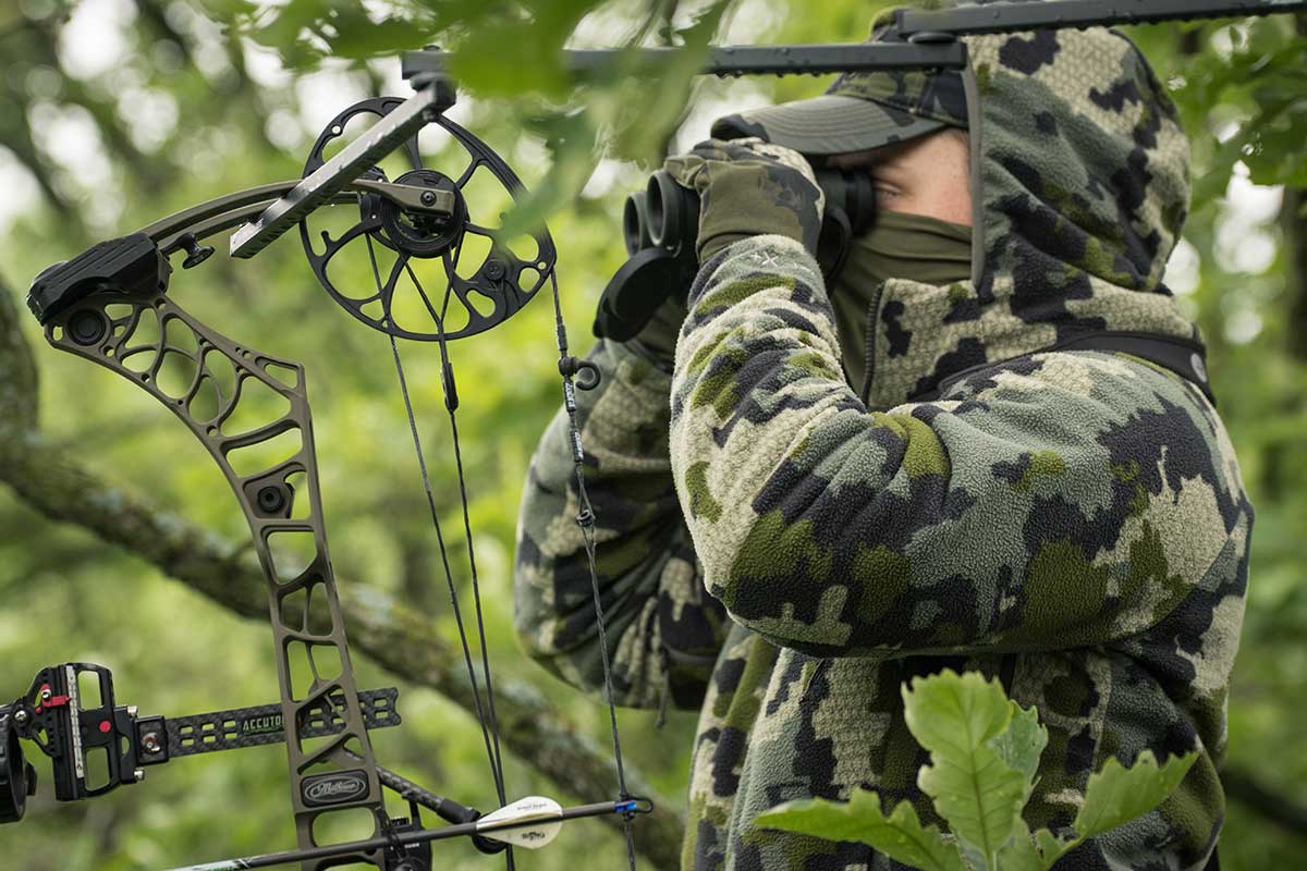 A hunter sits in a tree and looks through binoculars while wearing the new KUIU Proximity camo line.