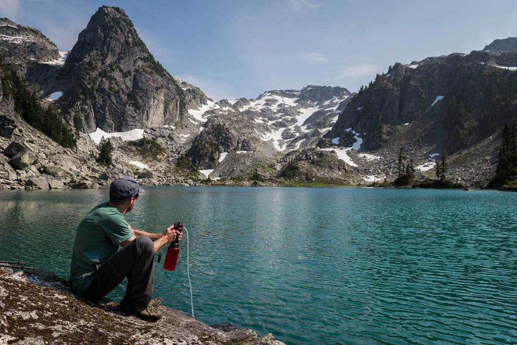 A man squats near a mountain lake to filter water for drinking.