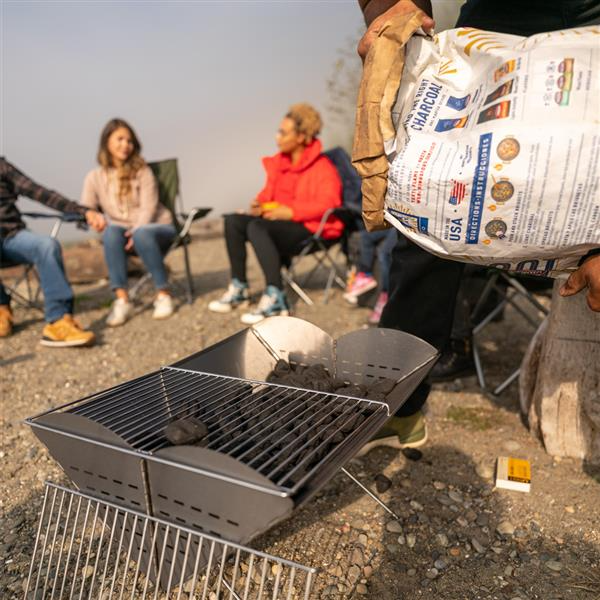 Charcoal getting poured into a UCO Flatpack Grill and Firepit with campers in the background.