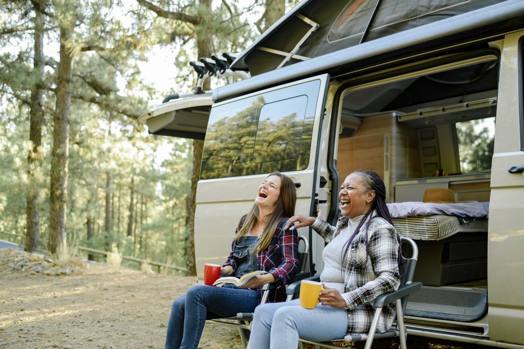 Excited diverse female friends sitting in camping chairs near traveling van and laughing at camping jokes while having fun in woods