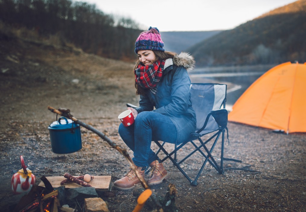 Alone at camping. One young woman preparing food at campfire next to the her tent and drinking hot tea on a cold autumn day