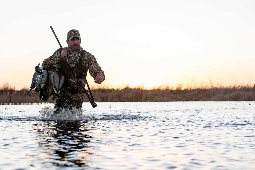 A duck hunter wades through water holding decoys and a shotgun.
