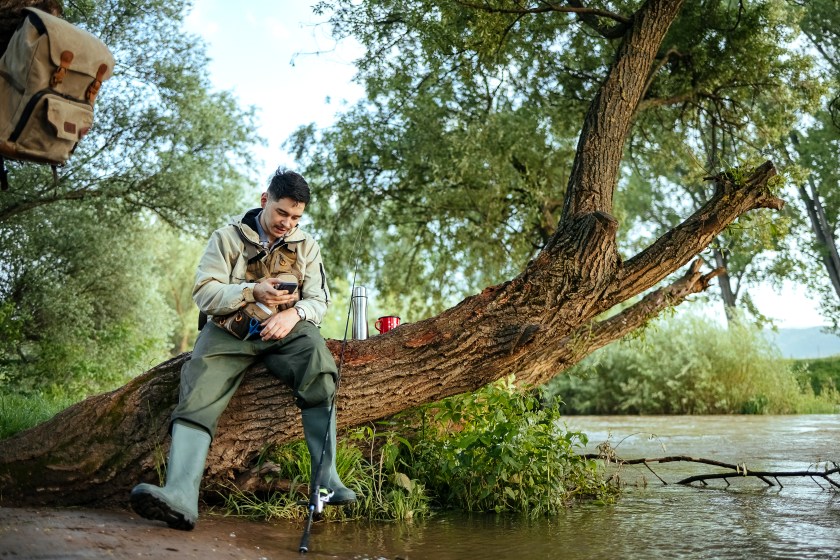 Shot of a young man using his cellphone while out fishing by a river