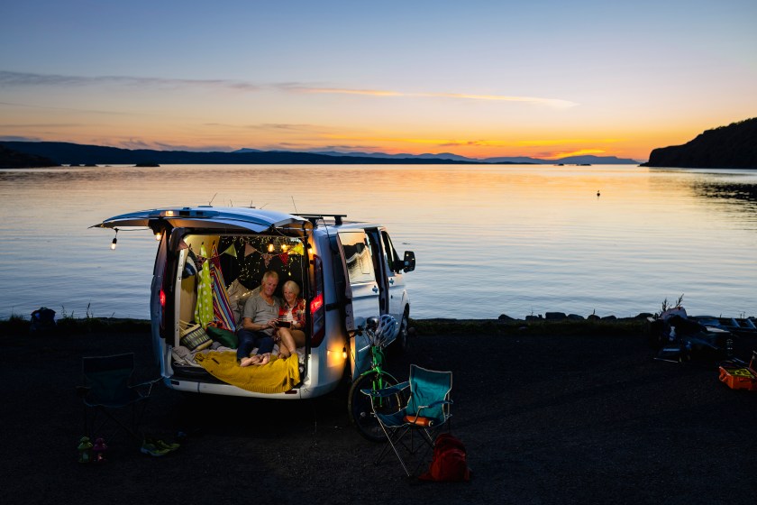 A high wide angle front view of a serene moment at sunset of a senior couple who are having a snuggle and watching something on a mobile phone in the back of their van which they have decorated with string lights and made comfortable with blankets. They are on the coast in Torridon in Scotland. 