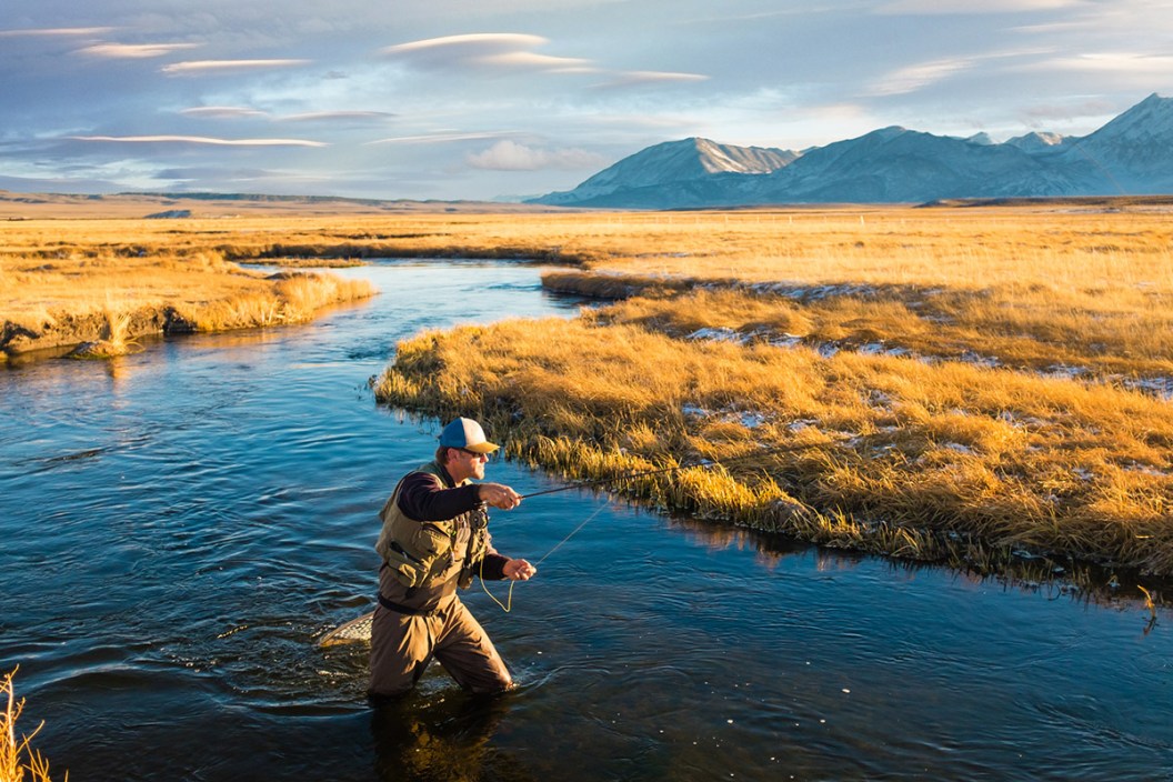 man in fly fishing gear on a river with the mountains behind