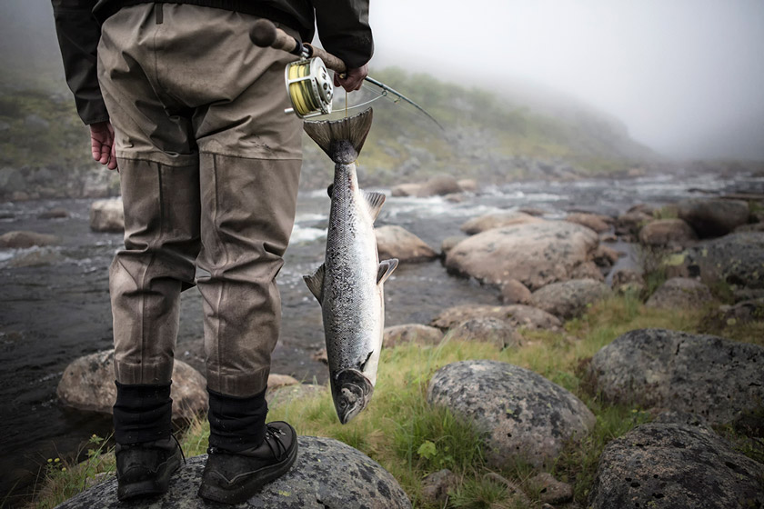 man in fly fishing waders holding fish on river bank