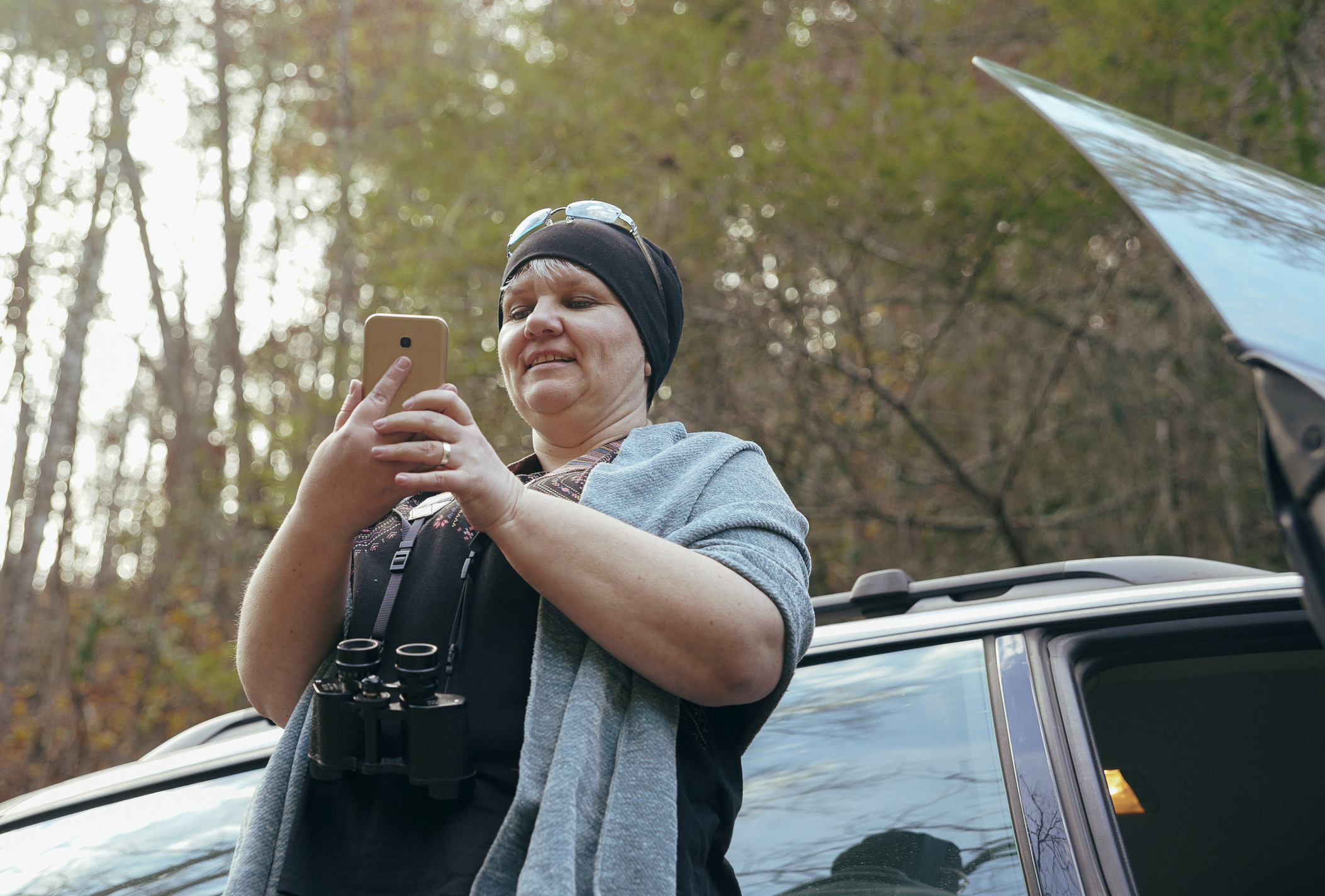 Woman uses phone in park while leaning against her car.