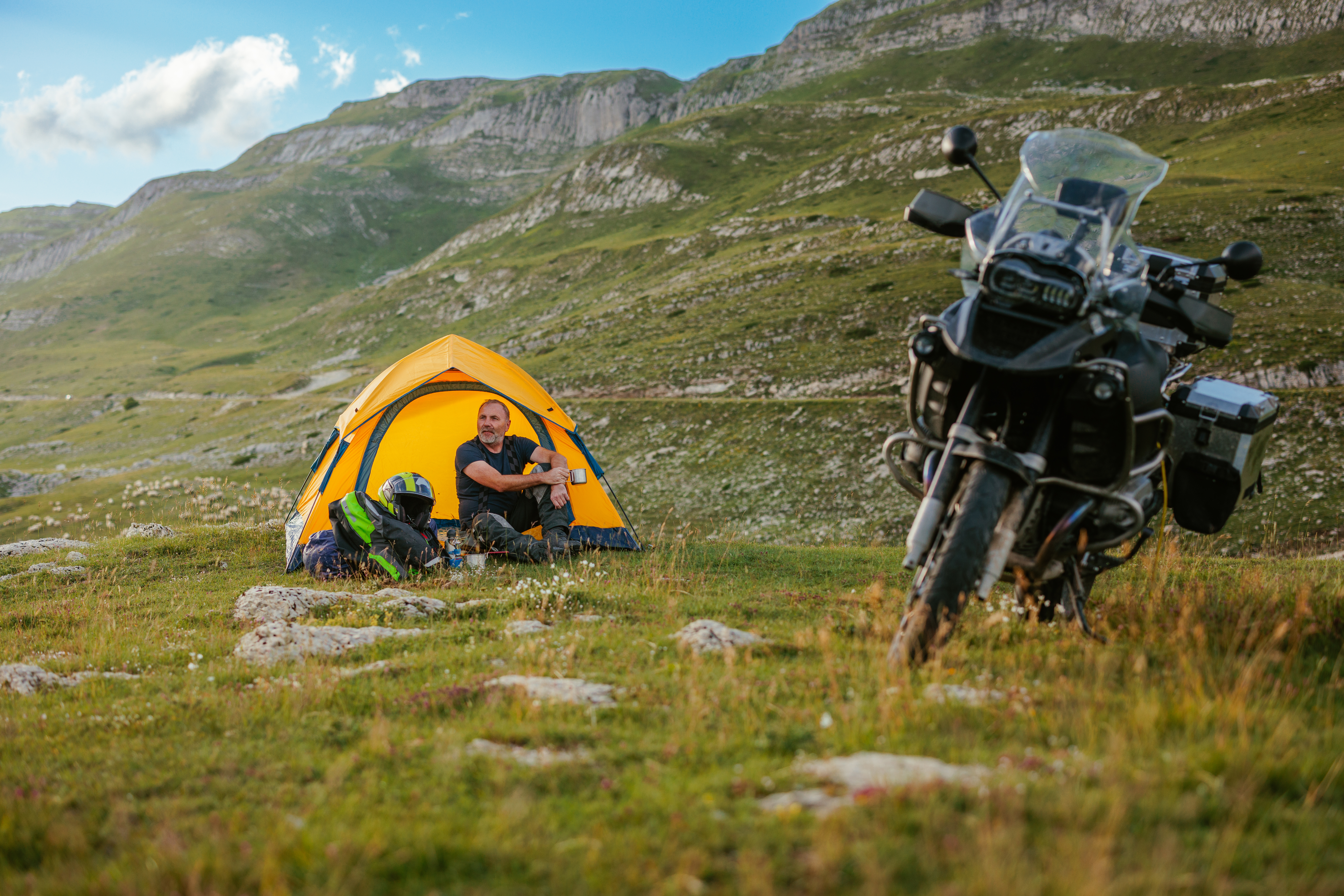 Biker rider is drinking coffee early in the morning in front of camping tent before hitting the road up in the mountains and marveling the landscape