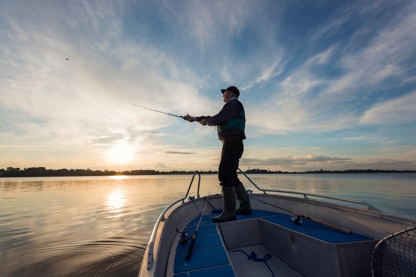 Portrait of a 65 year old fisherman casting out his fishing line into the water from his boat into a flat calm sea. Horizontal format photographed against a setting sun with lots of copy space in the sky. Photographed on Stege Nor on the island of Møn or Moen in Denmark.