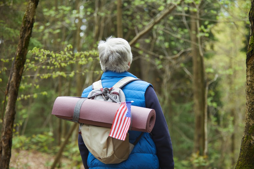 Back view at active senior man wearing backpack with American flag while enjoying hike in forest