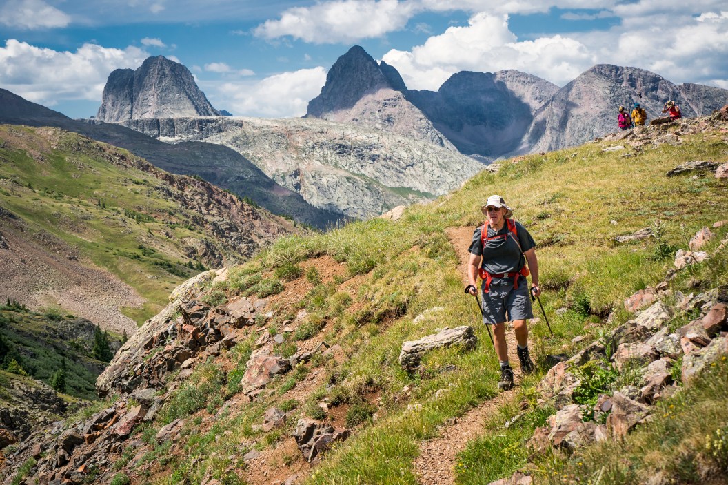 Adult man hikes a dirt path in a valley between dramatic mountain peaks on a sunny day, San Juan Mountains, Weminuche Wilderness, Rocky Mountains, Silverton, CO, USA