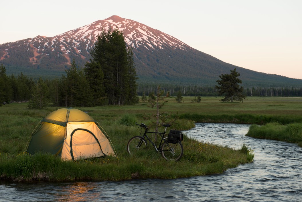 This image shows an alpine meadow w/illuminated tent after a day of road biking. Mt Bachelor in background. Pacific northwest cascades.