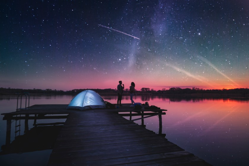 Couple camping on lake docks at dusk, standing next to a tent enjoying beautiful starry night sky
