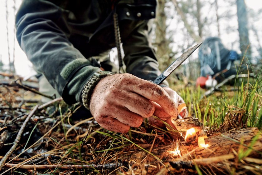 Close-up of man's hands igniting fire using ferrocerium rod and knife in forest.