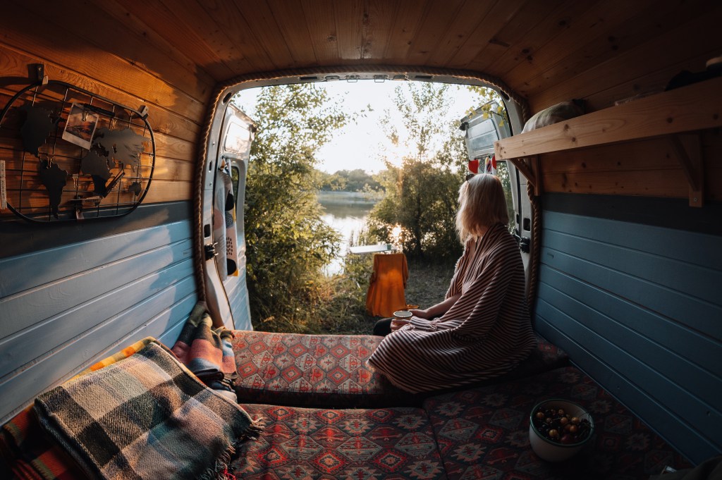 Woman living the van life experience watching a stunning view as she sits in a camper van and drinks coffee.