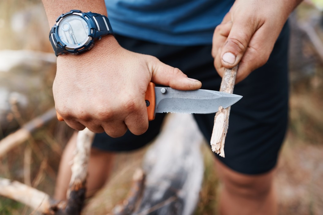 Hands, knife and cutting with a hiking man carving a stick outdoor in nature while camping for adventure. Wood, weapon and tool with a male camper slicing a twig in the mountains for survival