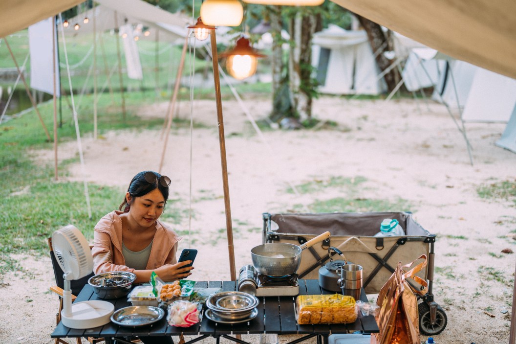 An Asian female using smartphone and prepare food on gas stove in campsite at forest