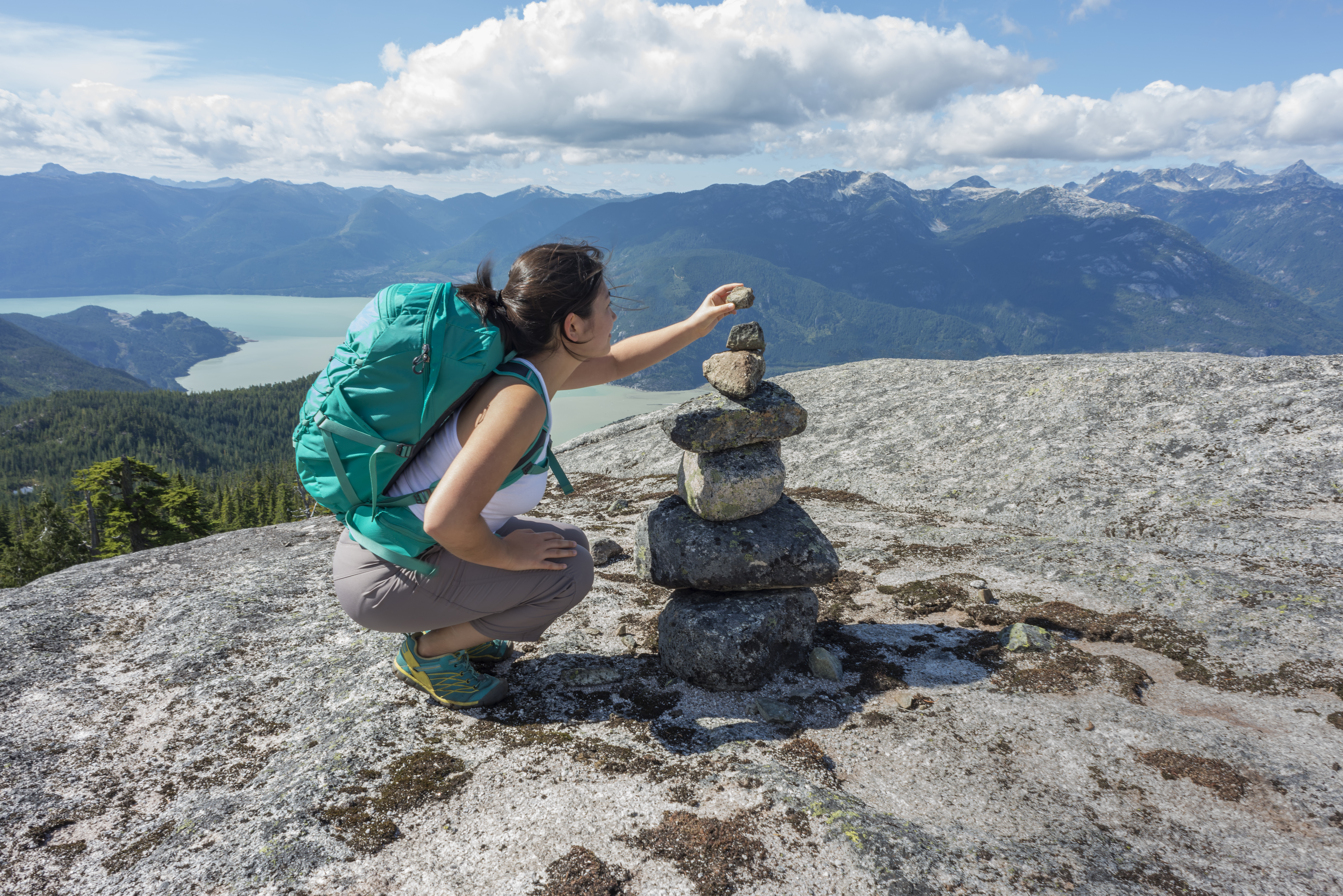 A young, female Asian backpacker adds a stone to a cairn on a mountaintop in Squamish, British Columbia, Canada. View of the valley and surrounding mountains in the background.