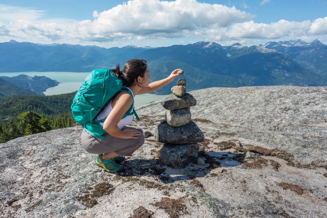 A young, female Asian backpacker adds a stone to a cairn on a mountaintop in Squamish, British Columbia, Canada. View of the valley and surrounding mountains in the background.