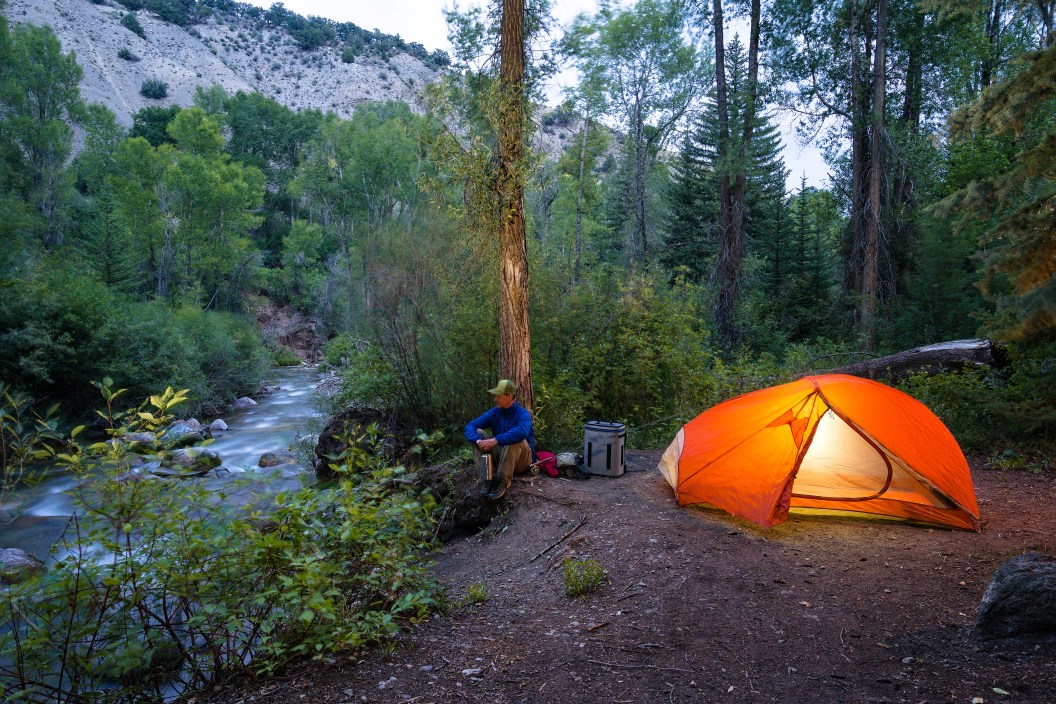 Fly Fish Backcountry Camp Along Creek - Tent and fishing gear with fly fisherman at dusk with glowing tent and flowing creek. Great outdoors adventure.