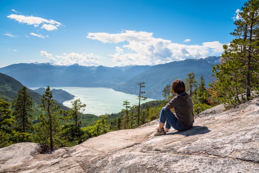 Woman Sitting Alone on a Rock on the Top of a Mountain admiring the Majestic Scenery with a Fjord Surrounded by Forested Coastal Mountains. Squamish, BC, Canada.