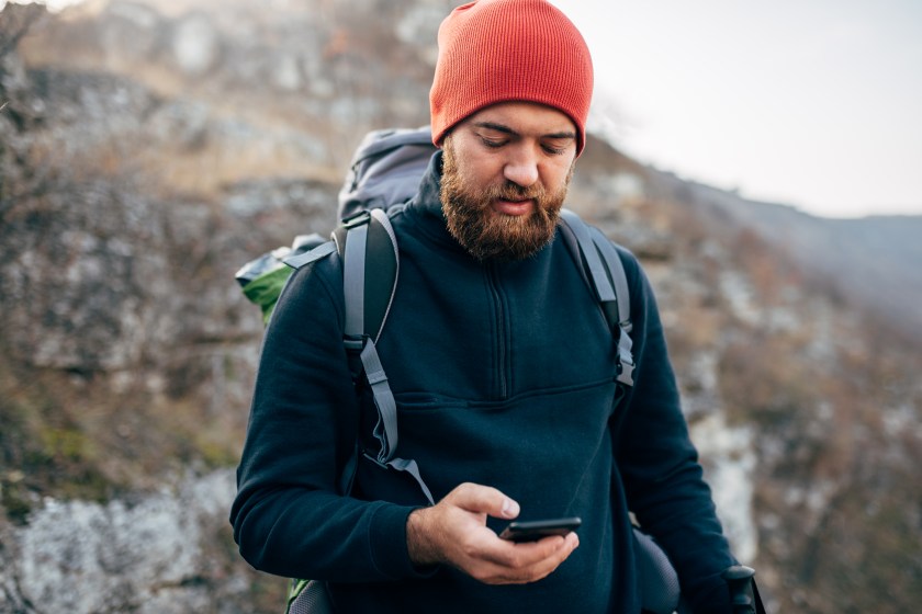 Portrait of young explorer bearded man trekking in mountains, using mobile phone for GPS application. Traveler man texting messages and browsing on Internet via his smart phone after hiking in mountain