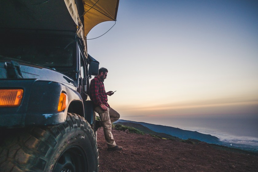 Traveler people with car and camping concept - lonely man use cellular phone to connect to internet outside his vehicle - mountain and nature outdoor around - enjoying freedom and alternative vacation with independence