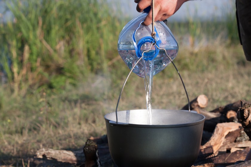 Adding water from a big plastic bottle in black cauldron on open fire, camping meal