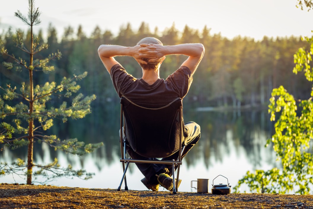 Man is sitting in a camping chair on the background of a forest lake on a beautiful summer evening. Object in focus, background blurred.