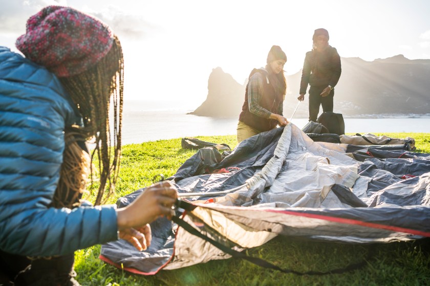 Diverse group of young friends lining up poles while setting up their tent overlooking the ocean during a camping trip together