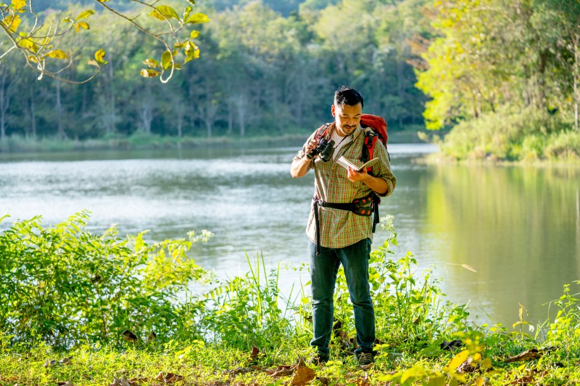 Asian man stand with look at book that record some data and he also hold binoculars look like he found some interesting creatures in the forest.