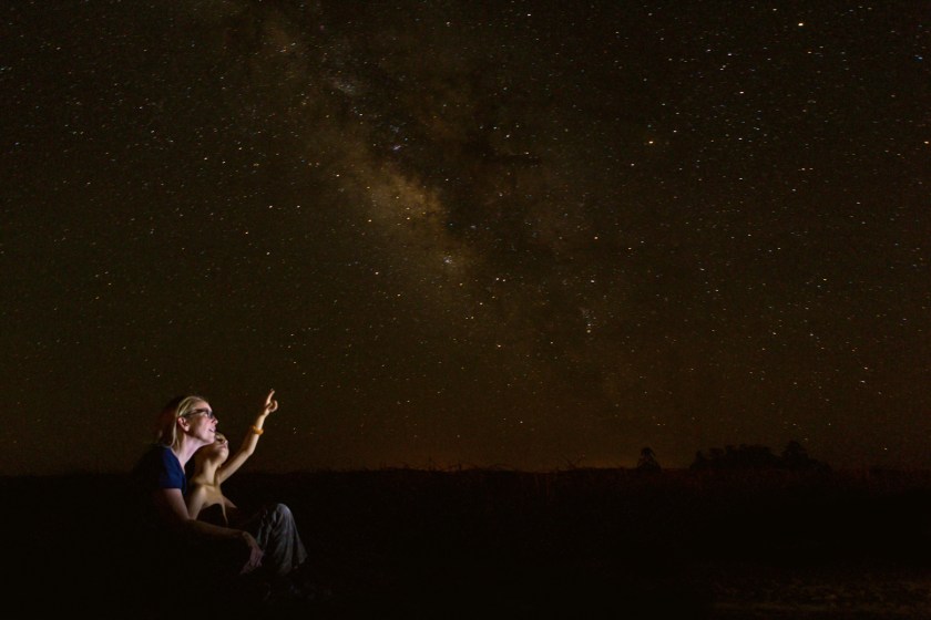 Mid adult Caucasian mother is sitting outdoors with her young son. Boy is sitting in mother's lap while they look up into the night sky. They are star gazing and studying constellations. Little boy is pointing to moon or stars in clear night sky.