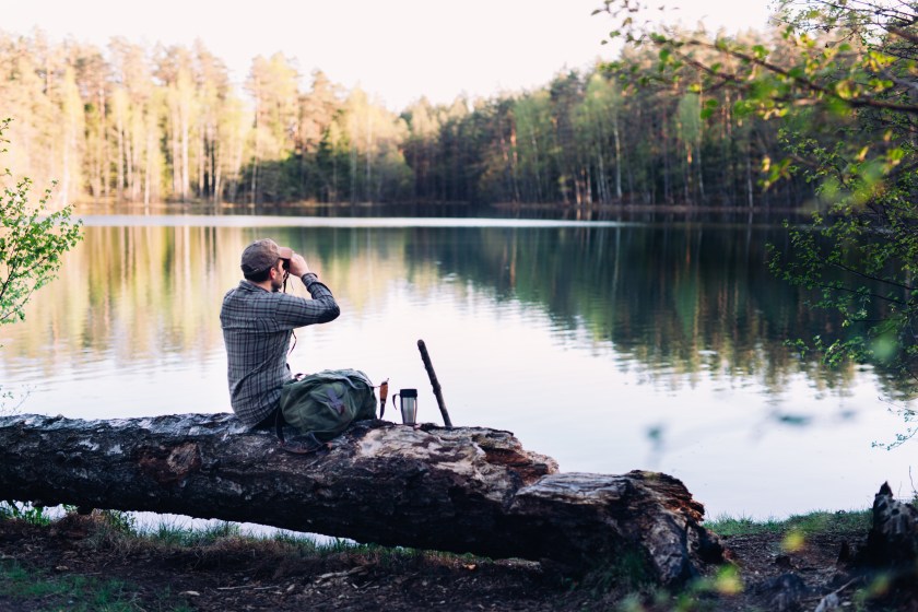 Mid aged hiker is sitting by the lake during sunset and observing the fauna using binoculars. He also has a backpack, pocket knife and a walking cane with him. Beautiful background with a sunlit forest and reflection on the lake. Guide to birding