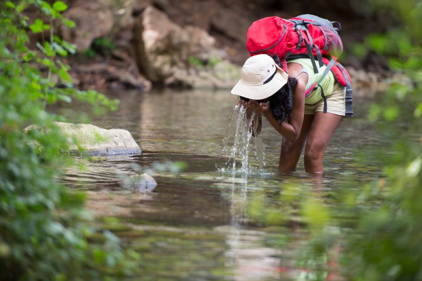 Backpacker tourist woman drinking and refreshing her face with the spring natural water from a mountain river. Woman wearing short shorts, tank top, fishing hat and red backpack.