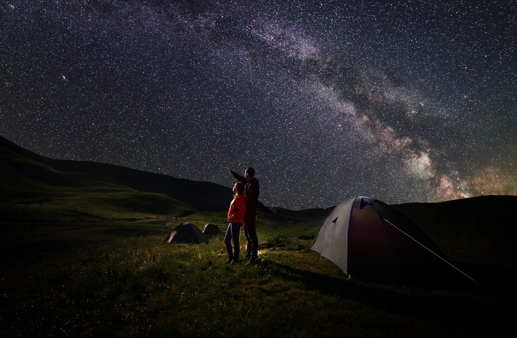 Young couple hikers standing near tents at night camping under starry sky against the backdrop of the mountains covered by greenery and lake. Guy pointing to the evening starry sky and Milky way