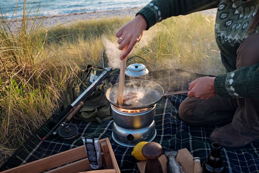 Fisherman cooking her catch at the beach. Female fisherman preparing a filled piece of fish she caught earlier. She is using a gas camping stove in a sheltered spot in the dunes next to the beach where she was fishing. Colour horizontal with some copy space. She is wearing a Nordic style sweater and hiking trousers. Shot on location on the island of Møn in Denmark.