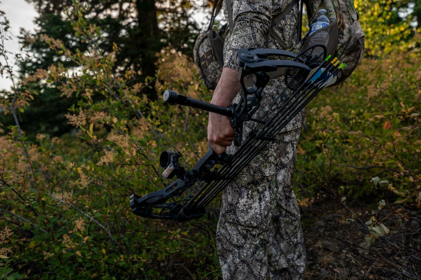 Close up shot of a hunter dressed in camouflage clothing holding a crossbow at his side while hunting elk in a forest at sunset. 
