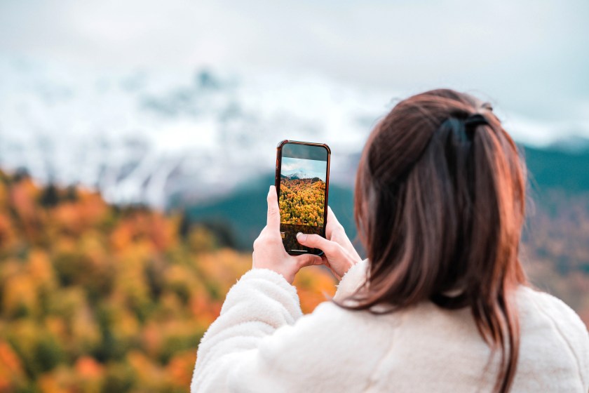 Happy tourist woman taking photo with smart phone on top of mountain in spring or autumn