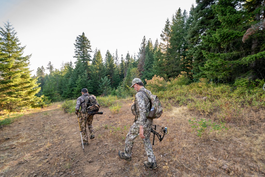 Two men wearing camouflage clothing and backpacks hike through a mountainous forest in Washington state while hunting elk early in the morning. One of the men is carrying a crossbow.
