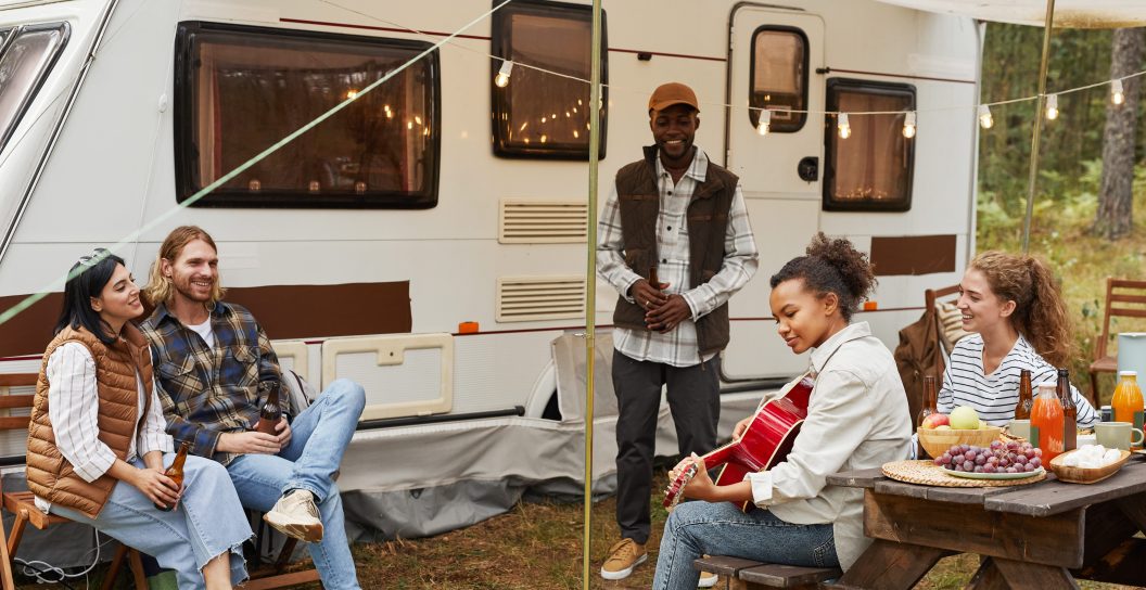 Full length portrait of young African-American woman playing guitar while enjoying camping outdoors with diverse group of friends, while RV boondocking.