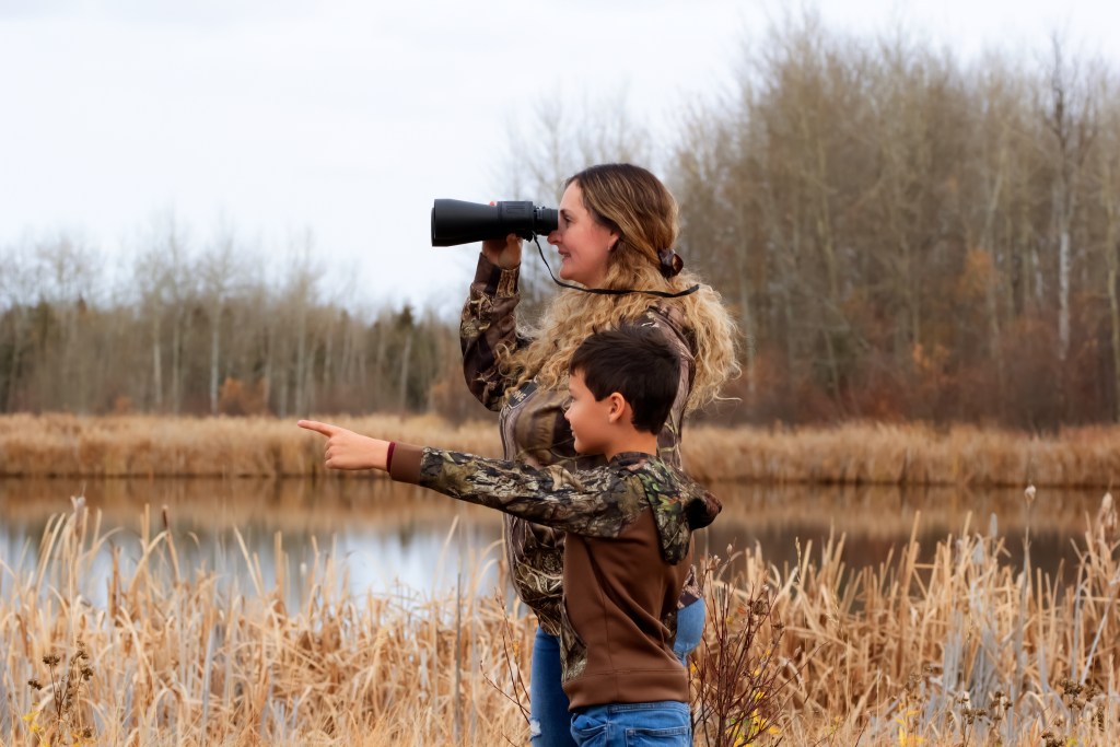 A mother and a son are in the park near the kale in late fall. Mother is watching in the binoculars and the boy is pointing at something.