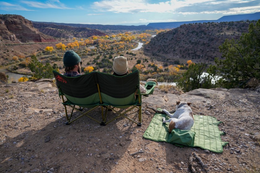 Two people sitting in a green camping loveseat, with a dog lying on a green mat next to them
