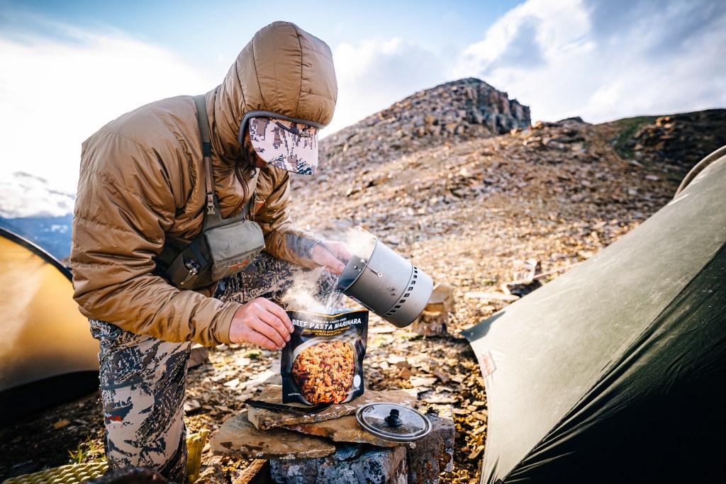 Person in a Sitka Jacket on a trail pouring a pot of boiling water into a bag of dried food