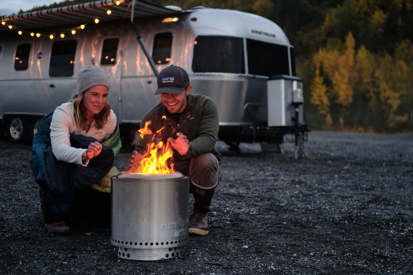 Two people sitting in front of a parked trailer behind a Solo Stove campfire