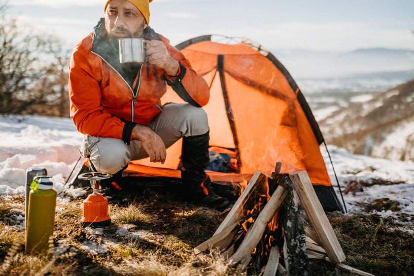 Mature man drinking hot drink in metal cup near tent on winter camping
