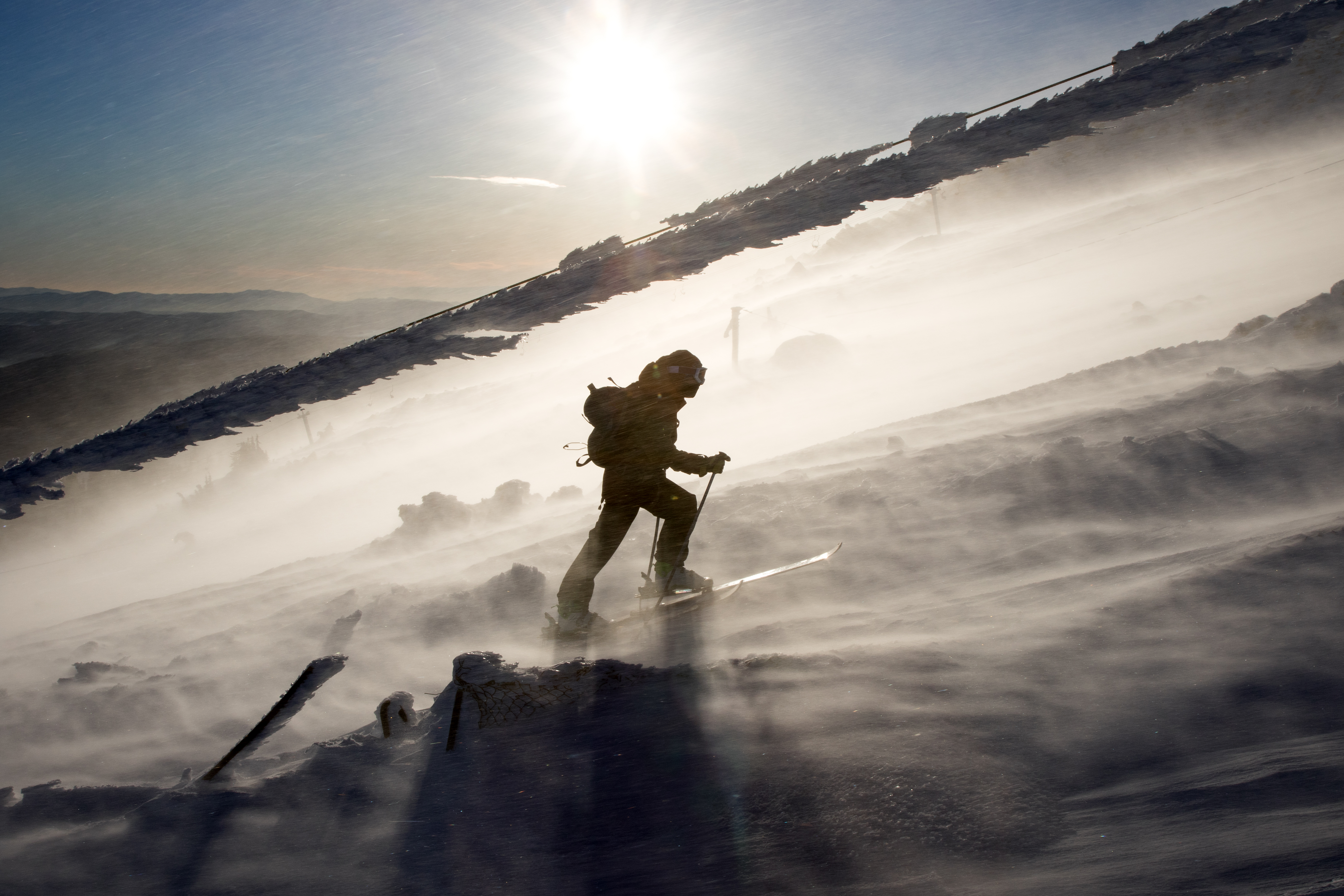 Back country skier walking up under a frozen cable of an old ski lift, in a severe windy weather.